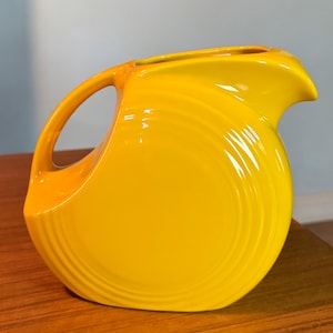 May include: A bright yellow ceramic pitcher with a curved handle and a spout. The pitcher has a glossy finish and a rounded body with subtle, vertical ridges. The handle is a lighter shade of yellow.