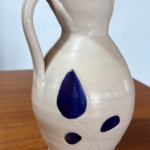May include: A light beige ceramic pitcher with a rounded body, a curved handle, and a narrow neck. The exterior features a hand-painted design of three dark blue teardrop-shaped leaves connected by thin, incised stems on the front surface.