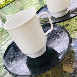 May include: Two white ceramic mugs with a ribbed design, sitting on black coasters with a white floral pattern.