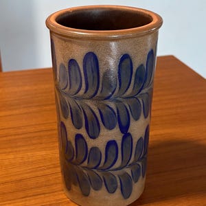 May include: A cylindrical ceramic vase with a brown exterior and a dark blue leaf pattern. The rim and interior are a darker brown. The vase is sitting on a wooden surface.