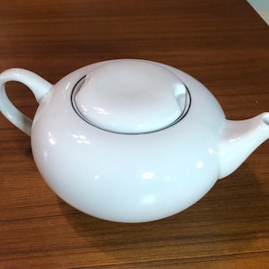 May include: A white ceramic teapot with a rounded body, a curved handle, and a spout. The lid has a black trim. The teapot is designed for brewing tea and has a classic, elegant appearance.