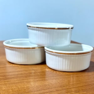May include: Three white ceramic ramekins with a gold rim. The ramekins are stacked on a wooden surface. Each ramekin has vertical ridges on the sides.
