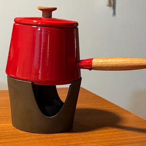May include: A red enameled fondue pot with a wooden handle and lid knob. The pot sits on a dark brown stand. The handle is approximately 6 inches long.