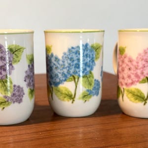 May include: Three white ceramic mugs with floral hydrangea designs in purple, blue, and pink. Each mug features a green rim and a matching floral pattern with green leaves. The mugs are cylindrical with a rounded base.