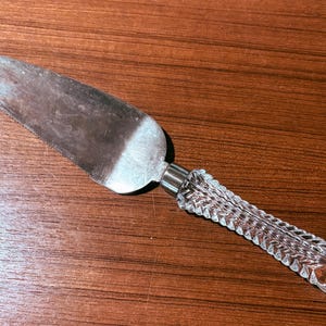 May include: A silver cake server with a serrated edge and a clear, textured glass handle. The handle has a decorative, ridged design. The server is resting on a brown wooden surface.