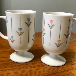 May include: Two white ceramic coffee mugs with a footed base. Both mugs have a pattern of stylized flowers in shades of blue, pink, orange, and green on a white background.