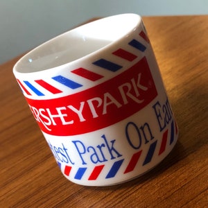 May include: White ceramic mug with the text "HersheyPark" in red and "Greatest Park On Earth" in blue. The mug has a red and blue striped design around the top.