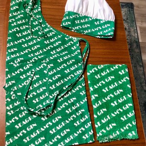 May include: A green kitchen set featuring an apron, chef's hat, and towel, all with a repeating white text pattern that reads "Seagram's Gin." The apron has a pocket and ties. The chef's hat is white with a green band. The towel has a fringed edge.