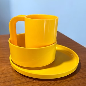May include: A bright yellow mug with a matching saucer and bowl. The mug has a large handle and sits inside the bowl. The set is made of a solid, smooth material and is placed on a wooden surface.
