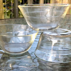 May include: Three clear glass bowls of varying sizes are stacked on top of each other. The bowls have a simple design with a few horizontal lines around the rim.