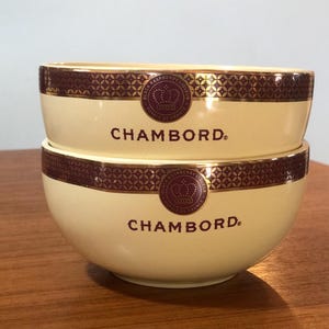 May include: Two cream-colored bowls with gold and burgundy trim. The bowls are stacked on top of each other. The top bowl has the text " Chambord" printed on it. The bottom bowl has the text " Chambord" printed on it.