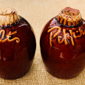 May include: Two brown ceramic salt and pepper shakers with cork tops. The salt shaker says "Salt" and the pepper shaker says "Pepper".