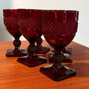 May include: Set of ruby red goblets. Each glass features a square base, a textured stem, and a diamond-patterned band around the top. The glasses are arranged on a wooden surface, showcasing their rich color and design.