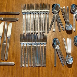 May include: A collection of stainless steel silverware, including knives, forks, spoons, and a cake server, arranged on a wooden surface. The silverware has a simple, elegant design, perfect for a formal dining setting.