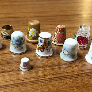 May include: A collection of ten thimbles, including a wooden thimble with a daisy design, a glass thimble with a red heart, and several porcelain thimbles with floral and butterfly designs.