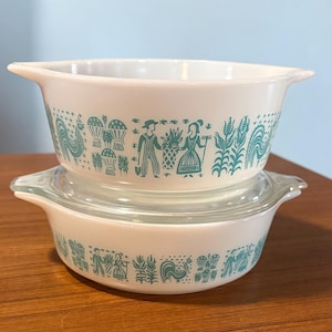 May include: Two white Pyrex bowls with a turquoise design featuring farmers, wheat, and roosters. The bowls are stacked, with one having a clear glass lid. The design wraps around the bowls.