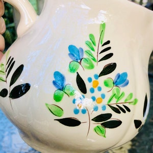 May include: Cream-colored ceramic pitcher with a curved handle and a wide spout. The pitcher is decorated with blue, green, and yellow floral designs. The flowers and leaves are hand-painted in a whimsical style.