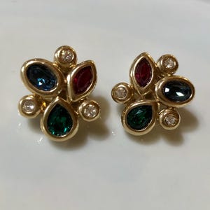 May include: A pair of gold tone earrings with a cluster of green, blue, and red gemstones. The earrings are set in a flower-like design.