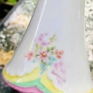 May include: White ceramic bell with a pink, yellow, and green trim. The bell has a floral design with pink, white, and blue flowers. The bell is marked "Sunrise, Inc."