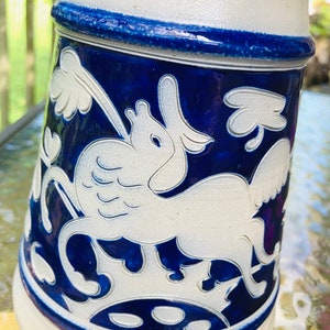 May include: A white ceramic stein with a blue and white design. The design features a stylised white bird with wings and a blue background. The stein has a blue rim and base.