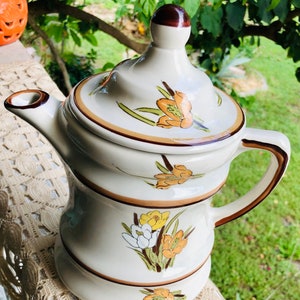 May include: A white ceramic teapot with a brown lid and handle. The teapot is decorated with a floral pattern in shades of orange, yellow, and white.