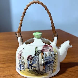May include: A small, white ceramic teapot with a painted village scene in blue, red, and yellow. It has a woven handle and a green knob on the lid. The teapot is approximately 4 inches tall.