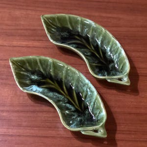 May include: Two green ceramic leaf-shaped serving dishes with a dark green and white glaze. The dishes are curved and have a raised edge.