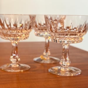 May include: Three pink-tinted crystal champagne coupe glasses. The glasses have a detailed, cut-glass design on the bowl and stem. The glasses are displayed on a wooden surface, with a neutral background.