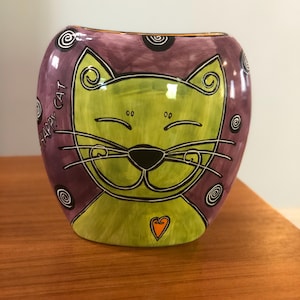 May include: A ceramic vase shaped like a cat with a green body, purple background, and black outlines. The cat has a happy expression with closed eyes and a small orange heart on its chest. The vase says "Happy Cat" on the side.