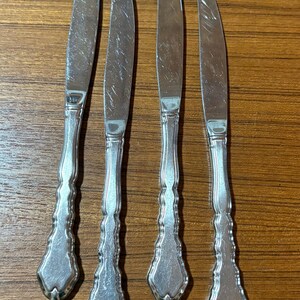 May include: Four silver-toned metal knives arranged vertically. Each knife features a smooth, pointed blade and a decorative handle with a series of vertical ridges and a pointed, shield-like base. The metal surfaces show visible surface scratches.