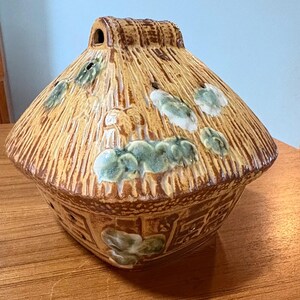 May include: A ceramic decorative house-shaped container with a thatched roof design. The roof is tan with brown accents and green leaf-like embellishments. The base has a textured pattern and additional green accents.
