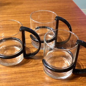 Op de afbeelding: Drie glazen bekers met zwarte kunststof handvatten. De bekers zijn leeg en staan op een houten oppervlak.