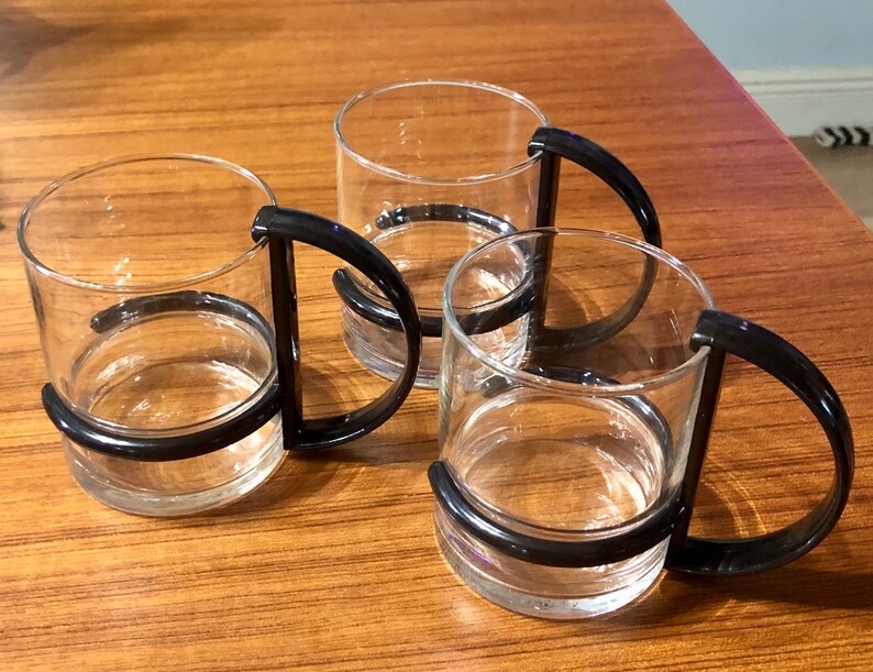 Op de afbeelding: Drie glazen bekers met zwarte kunststof handvatten. De bekers zijn leeg en staan op een houten oppervlak.