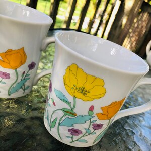 May include: Two white ceramic mugs with a floral design. The mugs feature yellow, orange, green, and purple flowers on a white background.