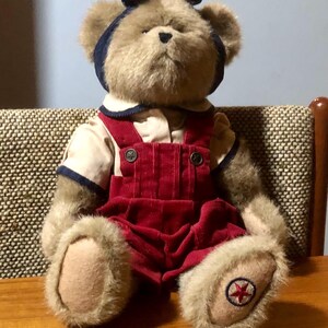 May include: A brown teddy bear wearing a red overall with a white shirt and a blue and white headband with a bow. The bear has a red star on the bottom of each foot.