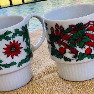 May include: Two white ceramic mugs with Christmas designs. One mug features a holly and poinsettia design, while the other has a design of bells and candles. Both mugs have a stacked design.