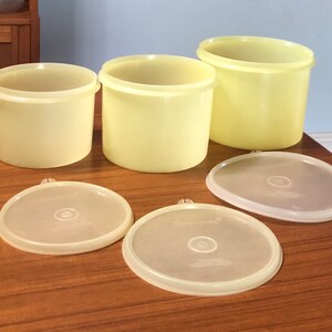May include: Set of three yellow plastic storage containers with lids. The containers are different sizes and are arranged on a wooden surface.