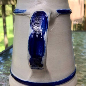 May include: A white ceramic beer stein with a blue handle and blue trim around the top and bottom. The stein has a traditional German design.