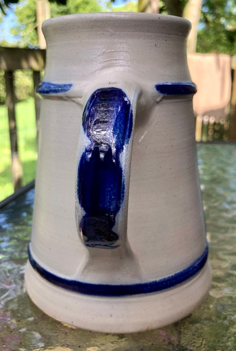 May include: A white ceramic beer stein with a blue handle and blue trim around the top and bottom. The stein has a traditional German design.