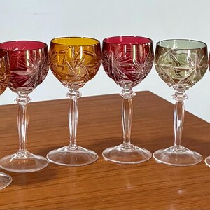 May include: Six stemmed wine glasses with colored bowls. The bowls are yellow, red, and green, with a starburst pattern. The stems and bases are clear. The glasses are arranged on a wooden surface.