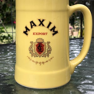 May include: A yellow ceramic beer stein with a large handle. The stein features the words "MAXIM EXPORT" in black lettering, along with a crest design in gold and red. The stein is sitting on a reflective surface.