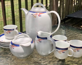 Op de afbeelding: Een wit porseleinen theeservies met blauwe en roze bloemmotieven. De set bevat een theepot, een roomkan, een suikerpot en vier kopjes en schoteltjes.