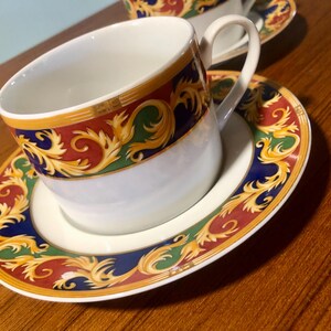 May include: A white teacup and saucer with a gold, red, green, and blue floral pattern. The cup has a handle and the saucer has a scalloped edge.