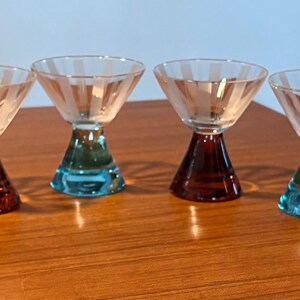 May include: Four vintage cocktail glasses with a conical bowl and a flared base. The bowls are clear with etched vertical lines, and the bases are in shades of brown and teal. The glasses are arranged on a wooden surface.