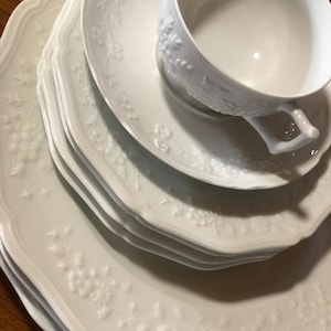 May include: A set of white porcelain plates and a teacup. The plates have a scalloped edge and embossed floral patterns. The teacup sits on a saucer, also with floral detailing. The set is clean and elegant.