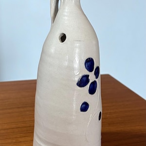May include: A tall, off-white ceramic jug with a narrow neck and a single side handle. The body features a small circular hole near the top and a dark blue floral design with five petals and a stem, embossed into the surface of the pottery.
