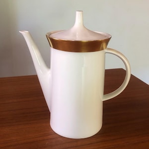 May include: White ceramic coffee pot with a gold trim around the lid and a gold band around the body. The pot has a spout and a handle.
