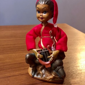 May include: A small, red-clad elf figurine playing a harp. The elf has a brown face, black hair, and a red hat with a gold trim. The harp is brown and has gold strings. The elf is sitting on a brown rock.