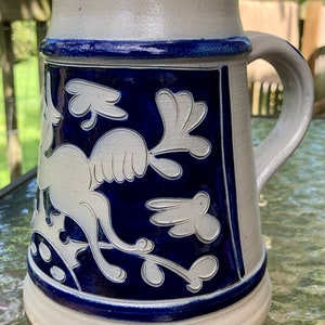 May include: A white ceramic beer stein with a blue glaze and a white design of a stylised bird with floral accents.