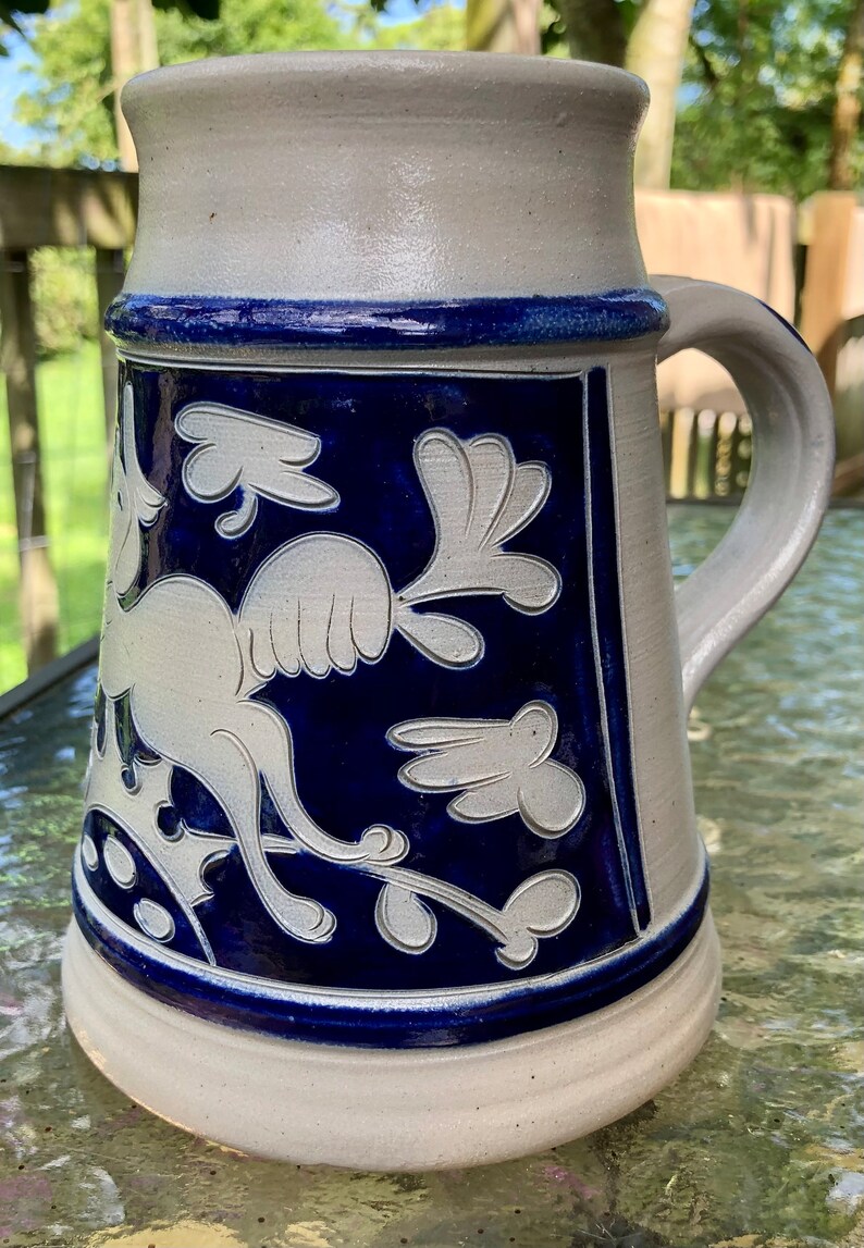 May include: A white ceramic beer stein with a blue glaze and a white design of a stylised bird with floral accents.